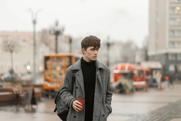 Stijlvolle moderne jongeman staat op een stadsmarkt met een heet café. Aantrekkelijk guy fashion model in modieuze winter buitenkleding loopt door de straat en drinkt een heerlijk drankje. — Stockfoto