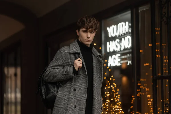 Un hombre joven con un peinado de moda en un abrigo de moda de temporada a cuadros gris con una mochila elegante en el hombro se encuentra en la calle cerca de un escaparate con luces brillantes. Guy modelo en la ciudad — Foto de Stock