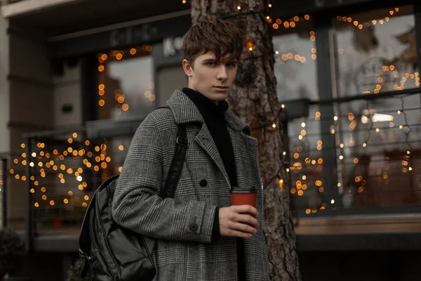 Urban ung man modell i eleganta vinter ytterkläder promenader längs gatan med varmt välsmakande kaffe nära vintage byggnader dekorerade med nyårsljus. Modern modekille går runt i staden. — Stockfoto