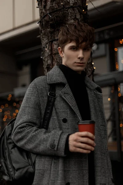 Joven guapo en un elegante abrigo a cuadros con una mochila de moda se levanta con una taza de delicioso café en la calle. Atractivo chico pasea por la ciudad en un fin de semana festivo con una bebida caliente . — Foto de Stock