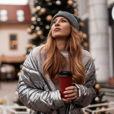 Bir fincan sıcak içecek standı ile şık kışlık giysiler içinde güzel bir kadının genç modeli ve oyuncaklarla eski bir Noel ağacının arka planına bakıyor. Kızıl saçlı kız şehirde dinleniyor.