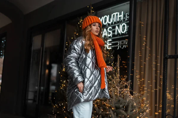 Red-haired pretty young woman in a fashionable knitted hat with an orange scarf in a trendy silver jacket poses outdoors near a window with Christmas decorations. Beautiful girl model. Time of magic. — Stock Photo, Image