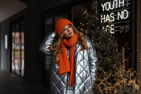 Mujer de pelo rojo joven europea en un elegante sombrero de punto con una bufanda naranja de moda en una chaqueta de plata de moda juvenil posando al aire libre cerca de una pared de vidrio con un árbol de Navidad y guirnaldas. Linda chica —  Fotos de Stock