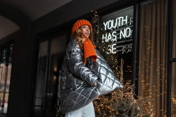 Aantrekkelijke mooie jonge vrouw in een stijlvol glanzend winterjasje met een warme gebreide muts met een oranje sjaal wandelt door de stad in de buurt van moderne gebouwen met bloemenslingers. Mooi rood haar meisje. — Stockfoto