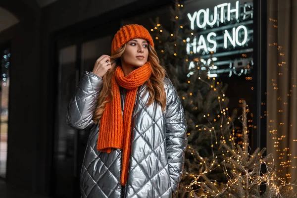 Gelukkige mooie jonge vrouw straalt haar. Modieus meisje in een stijlvol zilveren jasje in een oranje gebreide muts met een vintage warme sjaal poserend bij een kerstboom met lampjes op straat. — Stockfoto