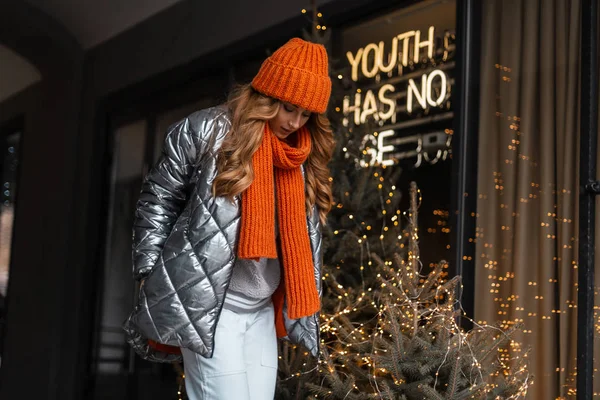 Trendy jonge vrouw met een warme hoed met een oranje gebreide sjaal in een glanzend modern jasje staat en kijkt neer bij een moderne glazen wand met vintage lichtjes. Meisje poseren in de stad op een warme winterdag. — Stockfoto