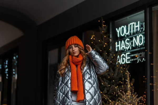 Mujer joven y bonita moderna con el pelo rojo en un sombrero de punto de moda con una bufanda naranja vintage en una chaqueta de plata brillante de moda juvenil posando en la ciudad cerca de una pared de vidrio con guirnaldas. Chica urbana . — Foto de Stock