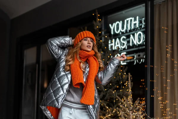 Modelo bastante joven de una mujer de moda en un elegante sombrero de punto naranja con una bufanda en una chaqueta brillante de moda juvenil posa en la ciudad cerca de un escaparate con luces brillantes.Muchacha de pelo rojo atractiva — Foto de Stock