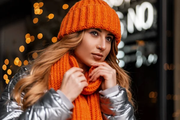 Roztomilá mladá žena v oranžovém klobouku s teplou pletenou šálou v mladém lesklém saku pózuje ve městě a sní o novoročním zázraku. Portrét krásné dívky v blízkosti výlohy obchodu se světly. — Stock fotografie