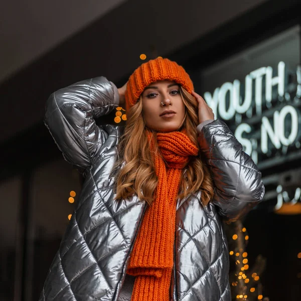 Jeune belle femme avec une coiffure dans un chapeau élégant tricoté avec une écharpe chaude orange dans une veste brillante à la mode posant dans la ville près d'un magasin moderne. Fille attrayante en vêtements d'hiver à la mode — Photo