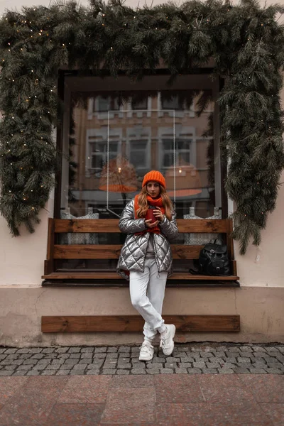 Stylish young woman with a cup of tasty hot drink is standing in Christmas weekend near the building with a vintage window. Urban girl in trendy winter glamorous clothes posing outdoors in the city. — Stock Photo, Image