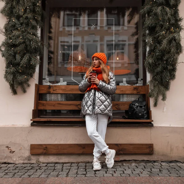 Europees model van een jonge vrouw in de winter stijlvolle buitenkleding staat met een kopje hete koffie in de buurt van een vintage venster met nieuwjaarsdecoraties. Aantrekkelijk meisje in trendy kleding wandelingen door de stad. — Stockfoto