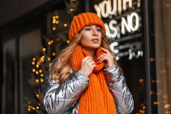 Bella donna urbana con un'acconciatura in un cappello elegante lavorato a maglia con una sciarpa calda arancione in una giacca lucida alla moda in posa in città vicino a un negozio moderno. Ragazza europea in outwear inverno alla moda . — Foto Stock