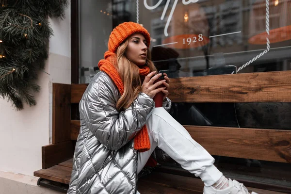 Die trendige junge Frau in winterlicher Oberbekleidung sitzt mit einer Tasse Kaffee auf einer Holzbank in der Nähe eines Vintage-Fensters auf der Straße. Nettes Mädchen, das sich draußen in der Stadt ausruht und einen heißen Tee trinkt. — Stockfoto