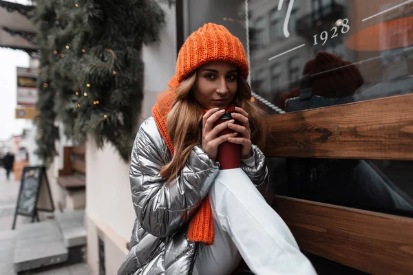 Современная молодая женщина в стильной зимней одежде в трикотажной шляпе с оранжевым шарфом с горячим кофе сидит на деревянной скамейке на улице у винтажного окна. Привлекательная девушка расслабляется на открытом воздухе . — стоковое фото