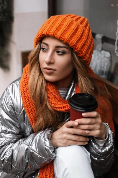 Retrato de una mujer joven con un sombrero naranja de moda con una bufanda en una chaqueta glamorosa brillante invierno cerca de una ventana vintage. Hermosa chica modelo se sienta con una taza de delicioso té y disfruta de un descanso . — Foto de Stock