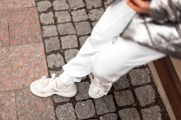 Top view on female legs in fashionable leather sneakers in trendy white pants outdoors. Urban girl in a glamorous white silver jacket in vintage shoes stands on a stone tile in the street. Close-up. — 스톡 사진