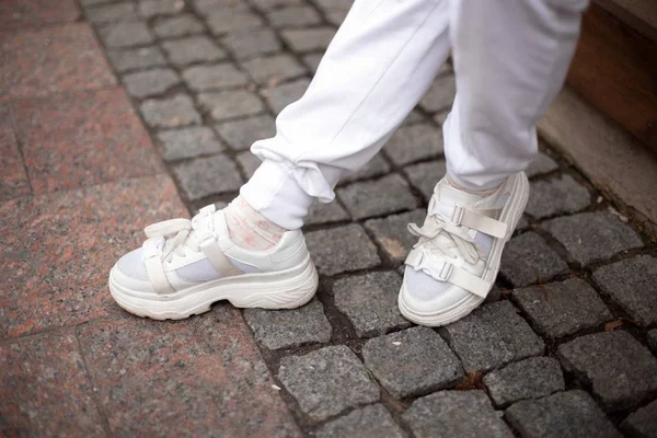 Närbild av kvinnliga ben i snygga läder vita sneakers i trendiga vita byxor på gatan. Modern ung kvinna står på en sten kakel på gatan i trendiga kläder. Ungdoms avslappnade stil. — Stockfoto