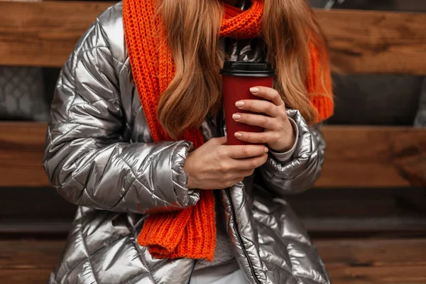 Primo piano di una giovane donna in una giacca alla moda lucida con una sciarpa arancione lavorata a maglia con una tazza rossa con caffè in mano su una panchina di legno in città. Ragazza elegante si rilassa e bere tè all'aperto . — Foto Stock