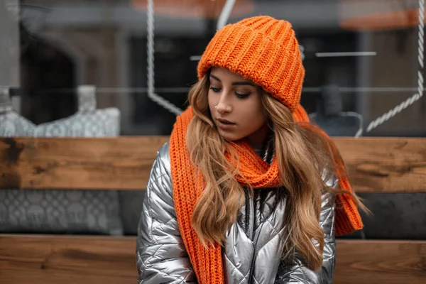 Bonita mujer joven con el pelo rojo en una chaqueta brillante de moda en un sombrero de punto naranja con una bufanda elegante se sienta y mira hacia abajo cerca de una ventana en la ciudad. Atractivo modelo de chica de moda . — Foto de Stock