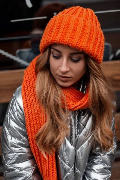 Atraktivní hezká mladá žena ve stylovém oranžovém pleteném klobouku s módní teplou šálu ve stříbrném retro bundě shlíží dolů venku.Krásná dívka model má relaxační venku v zimním dni — Stock fotografie