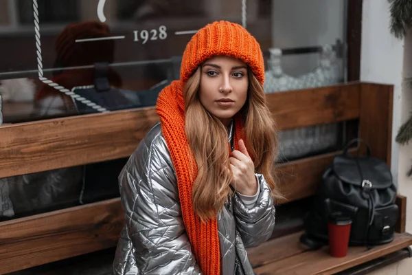 Европейская милая модель молодой женщины в модной вязаной шляпе в стильной блестящей куртке с винтажным шарфом сидит на деревянной скамейке возле стеклянной стены. Рыжеволосая девушка отдыхает в городе . — стоковое фото