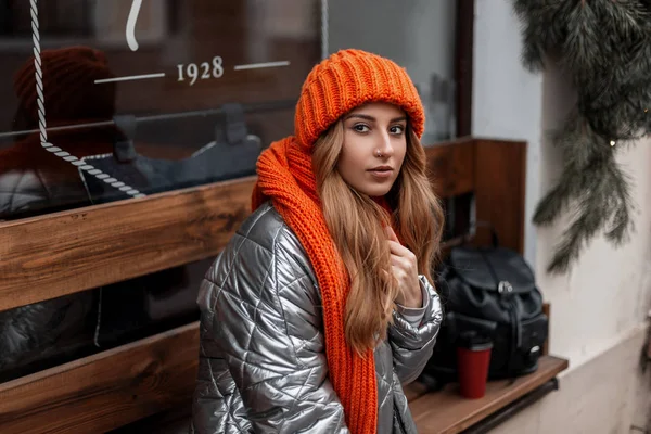 Young stylish woman in trendy warm winter clothes in a fashion knitted orange hat and scarf enjoys relaxes outdoors near a vintage wall in the street. Cute girl model sits on a wooden bench outdoors. — 스톡 사진