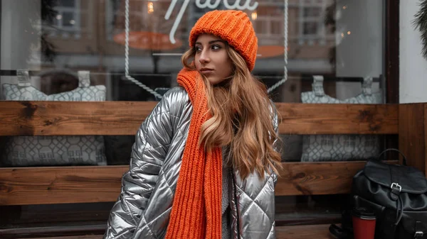 Modieuze jonge vrouw model in een stijlvolle gebreide hoed met een vintage warme sjaal in een trendy zilveren jas poseren op een bank in de buurt van een modern gebouw. Aantrekkelijk meisje in de stad. Jongerenstijl. — Stockfoto