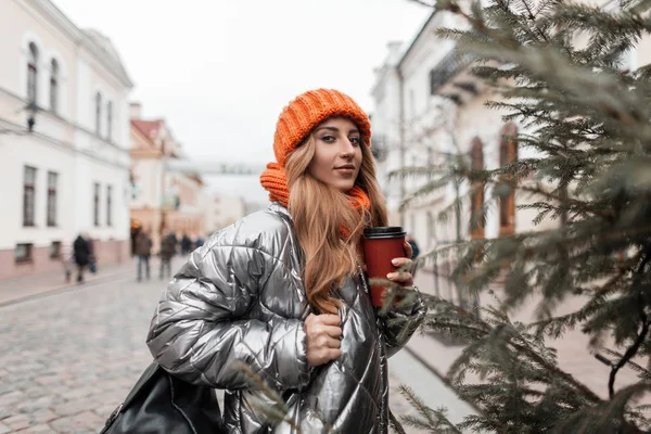 Весела красива молода жінка в модному зимовому срібному піджаку в в'язаному капелюсі з помаранчевим шарфом зі смачною кавою в руках насолоджується прогулянкою по місту. Щаслива дівчина в юнацькому верхньому одязі на вулиці — стокове фото