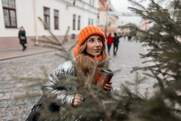 Happy young woman with a smile in stylish winter outerwear with a cup of hot drink walks on the street near the Christmas tree. Beautiful red-haired girl in trendy clothes is enjoys weekend outdoors.