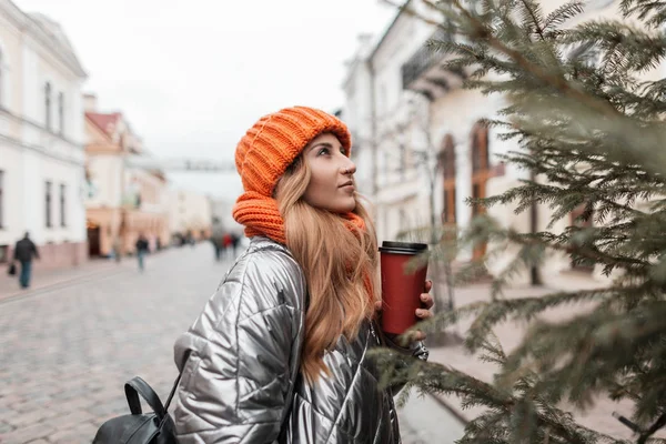 Trendy model van een schattige jonge vrouw in modieuze jeugd winterkleding met een leren rugzak met een kopje warme thee genieten van een wandeling in de stad. Stijlvol meisje staat en kijkt naar de kerstboom. — Stockfoto