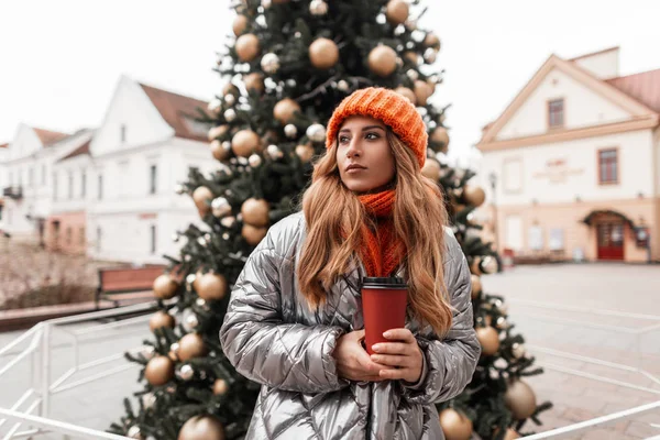 La giovane donna dai capelli rossi in un cappello arancione alla moda con una sciarpa lavorata a maglia in una giacca elegante con una tazza di tè in mani cammina sulla città vicino a un albero di Natale con giocattoli. Ragazza gode il fine settimana di Capodanno . — Foto Stock