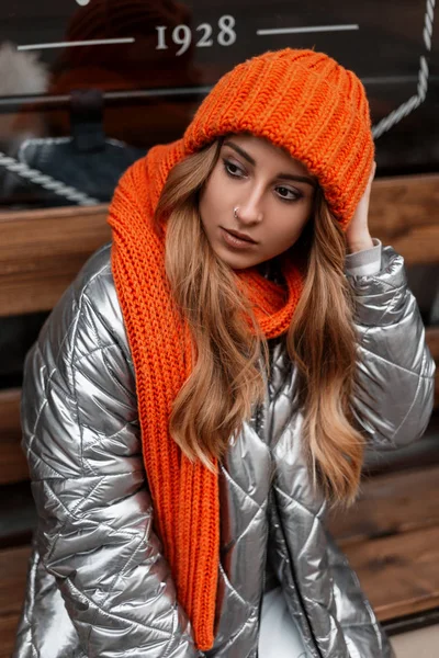 Jovem de cabelos vermelhos europeu em um chapéu de malha laranja na moda em uma jaqueta de inverno elegante posando perto de uma parede de vidro na cidade. Menina bonito em roupas da moda posando na rua. Olhar casual . — Fotografia de Stock