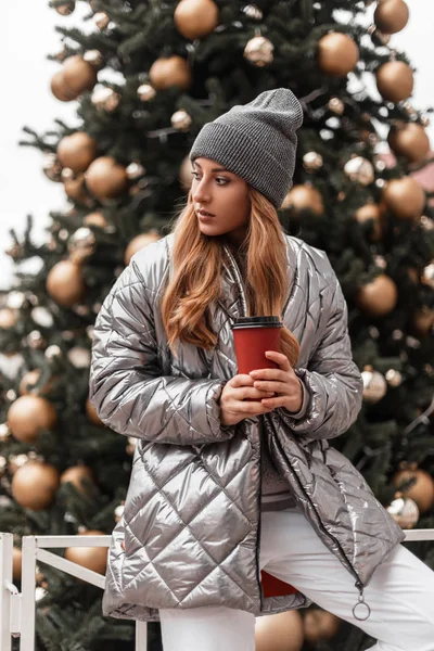 Eine junge Frau mit einer stylischen Strickmütze in einer modischen silbernen Jacke steht mit einer Tasse heißem Tee in der Hand neben einem schönen Neujahrsbaum mit Vintage-Spielzeug. hübsche trendige Mädchen ruht im Freien. — Stockfoto