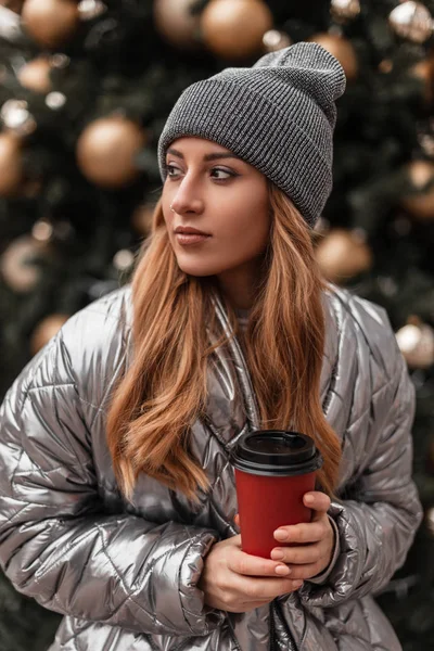 Retrato de uma jovem ruiva em um chapéu cinza de malha vintage em uma jaqueta de prata elegante com uma xícara de café nas mãos contra o fundo de uma árvore de Natal vintage festiva com brinquedos . — Fotografia de Stock