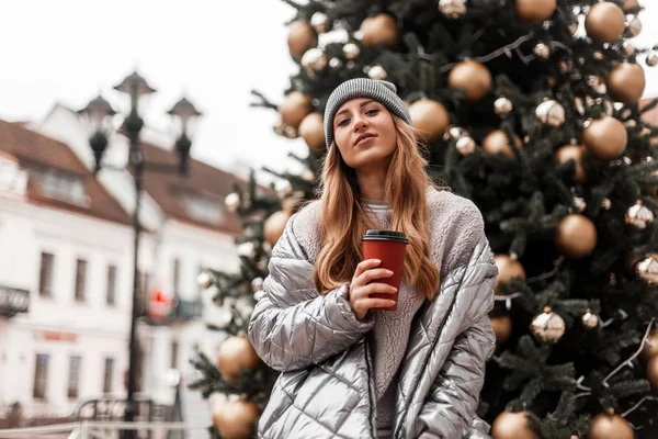 Весела модель молодої жінки в в'язаному сірому стильному капелюсі в срібній зимовій куртці стоїть з чашкою смачної кави в місті біля новорічної ялинки з золотими іграшками. Щаслива дівчина з милою посмішкою . — стокове фото