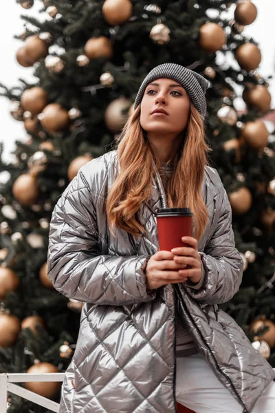 Jonge mooie vrouw met rood haar in een modieuze gebreide grijze hoed in een jeugd stijlvolle zilveren jas met een kopje koffie poses in de buurt van een vintage kerstboom met gouden speelgoed en feestelijke slingers. — Stockfoto