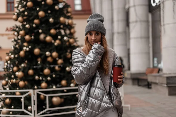 Giovane donna alla moda in un cappello elegante in una giacca d'argento giovanile con una tazza di bevanda calda posa vicino a un bellissimo albero di Natale festivo all'aperto. Moderna ragazza alla moda modella cammina per strada . — Foto Stock