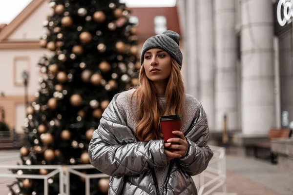 Міська стильна молода жінка в блискучій модній куртці в в'язаному модному капелюсі стоїть з гарячою кавою біля старовинної ялинки з золотими скляними кульками. Сучасна дівчина насолоджується прогулянкою в зимовий день . — стокове фото