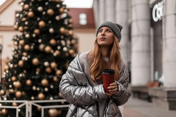 Young attractive woman with tasty latte poses on the background of the New Year tree in the street.Pretty beautiful girl fashion model in fashionable youth winter clothes enjoys a vacation in the city — Stock Photo, Image