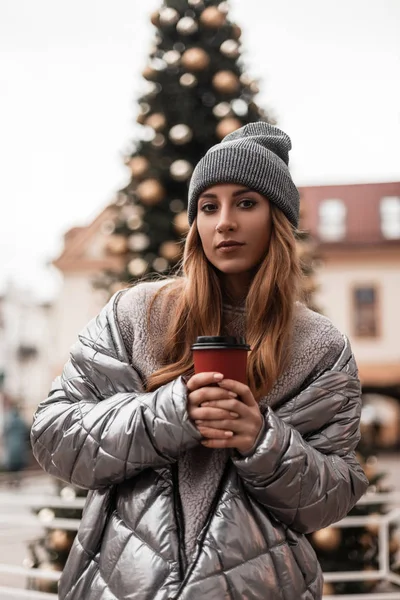 A jovem europeia com o cabelo vermelho em um chapéu tricotado na moda em uma jaqueta de prata elegante com uma xícara de chá em mãos está perto de uma árvore de Natal em uma rua na cidade. Menina da moda glamourosa . — Fotografia de Stock