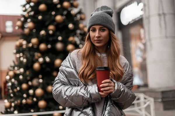 Vrij moderne jonge vrouw model in een gebreide grijze stijlvolle hoed in een glanzende winterjas staat met een kopje lekkere koffie in de stad in de buurt van een nieuwjaarsboom met gouden speelgoed. Schattig meisje. Jongerenmode. — Stockfoto