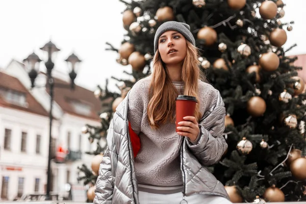 Mooie stedelijke jonge vrouw in een stijlvolle gebreide hoed in een modieuze zilveren jas staat met een kopje warme thee in de handen in de buurt van een mooie Nieuwjaarsboom met vintage speelgoed. Trendy meisje loopt op straat — Stockfoto