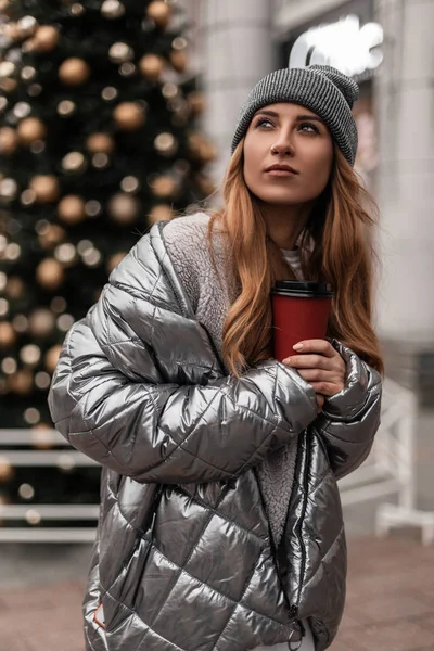 Attractive young woman in a stylish knitted hat in a fashionable silver jacket stands with a cup of hot tea in hands near a beautiful New Year tree with holiday toys. Cute girl walks around the city. — Stock Photo, Image