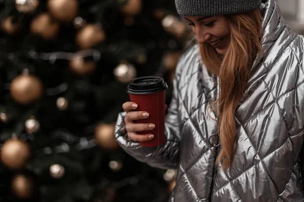 A jovem ruiva positiva em um chapéu de malha cinza em uma jaqueta elegante com uma xícara de café em mãos ri e olha para baixo no fundo de uma bela árvore de Ano Novo. Menina feliz atraente . — Fotografia de Stock