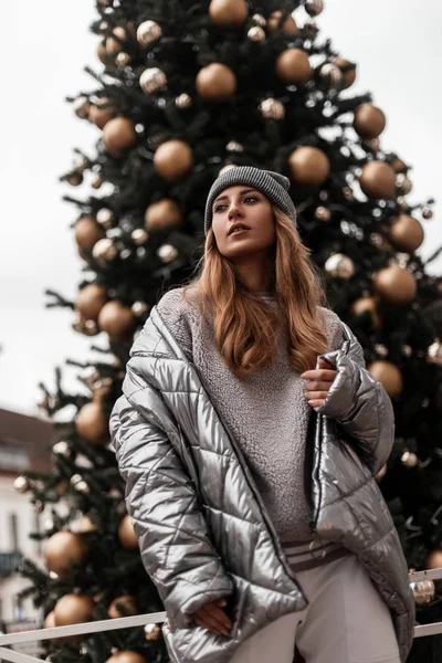 Model van een aantrekkelijke jonge vrouw in een grijze stijlvolle gebreide muts in een modieus zilveren jasje staat bij een prachtige nieuwjaarsboom met vakantiespeelgoed. Modern meisje loopt rond in de stad. Jongerenstijl. — Stockfoto