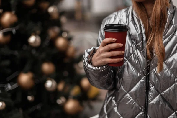 Close-up de uma xícara vermelha com chá saboroso em mãos femininas perto de uma árvore de Natal festiva com brinquedos vintage na cidade. Menina ruiva em uma jaqueta brilhante na moda caminha pela rua em um dia de inverno . — Fotografia de Stock