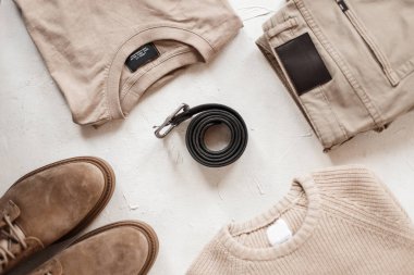 Deri siyah erkek kemerinin üst görüntüsü beyaz bir masada klasik tokalı sıradan moda kıyafetleri ve bej renkli ayakkabılar arasında. Erkekler için moda bahar koleksiyonu. Klasik bir tarz. Yakın plan..