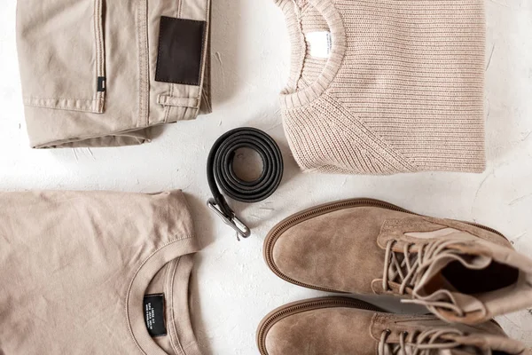 Černý stylový kožený pásek leží vedle pleteného svetru s klasickými kalhotami se semišovými botami a bavlněným svetrem na bílém stole. Pohled shora. Módní pánské oblečení v béžových barvách. — Stock fotografie