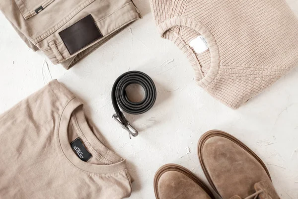 Fashionable black leather belt with a metal buckle on a table among fashionable men's clothing in beige colors. Closeup of a sweater with pants with a pullover with boots with a belt on a white table. — ストック写真